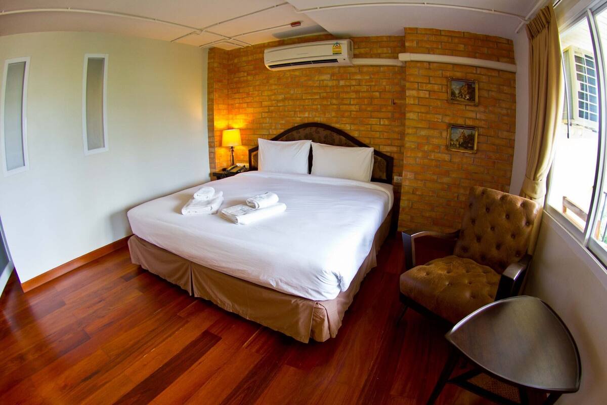 Habitación de hotel con una cama grande, suelo de madera, pared de ladrillo decorativa y una ventana con cortinas, además de una silla.