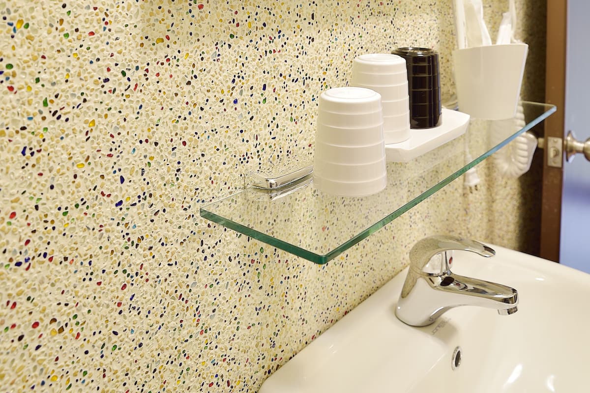 Une salle de bain avec un mur recouvert de carreaux à motifs, une étagère en verre où sont posés les articles de toilette, un lavabo avec un robinet chromé et un miroir qui reflète la pièce.