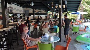 An outdoor seating area with round tables and chairs, a poolside area with lounge chairs, and a bar with a stone wall backdrop. - Club Turquoise Apartments (Marmaris)