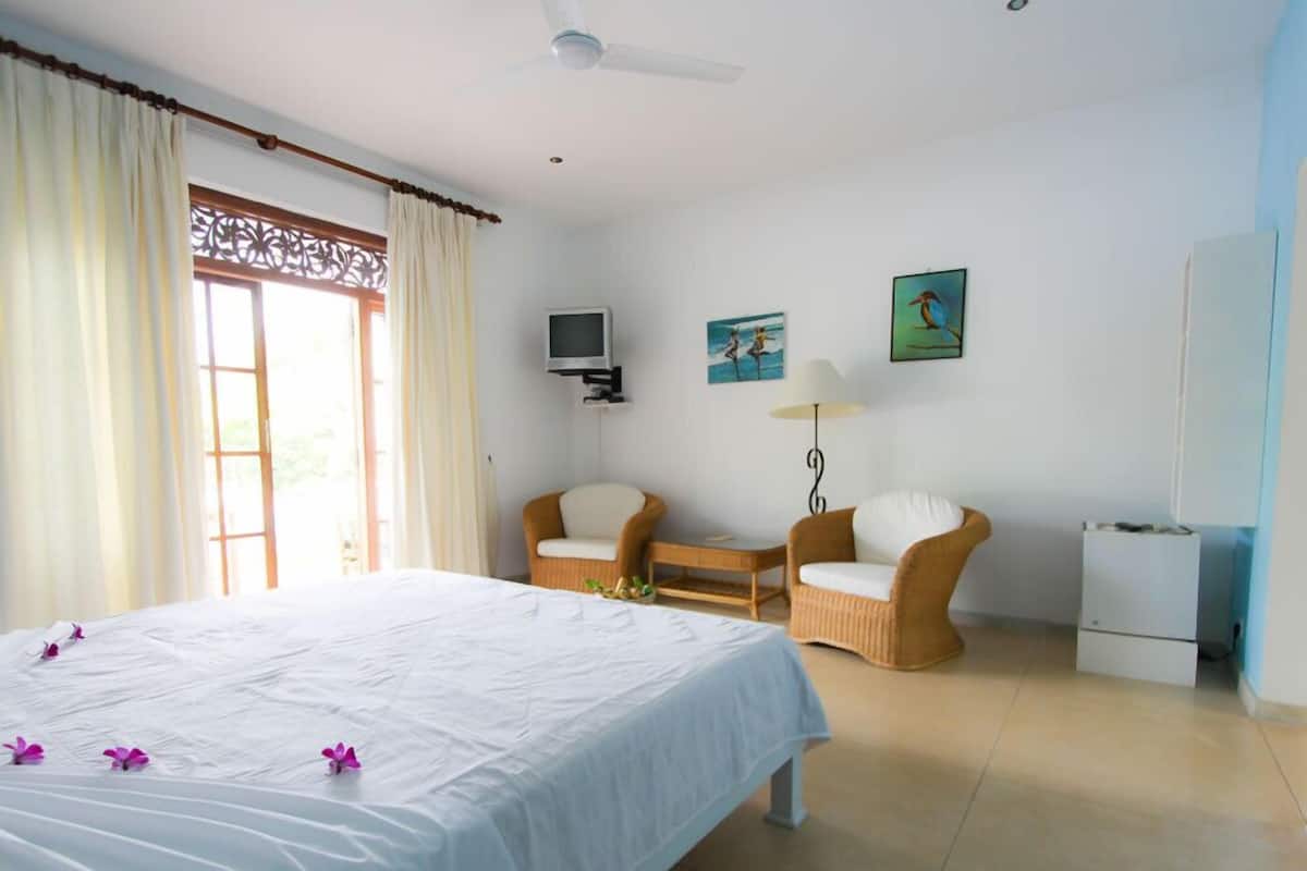 A hotel room with a bed, two chairs, a TV, and artwork on the wall.