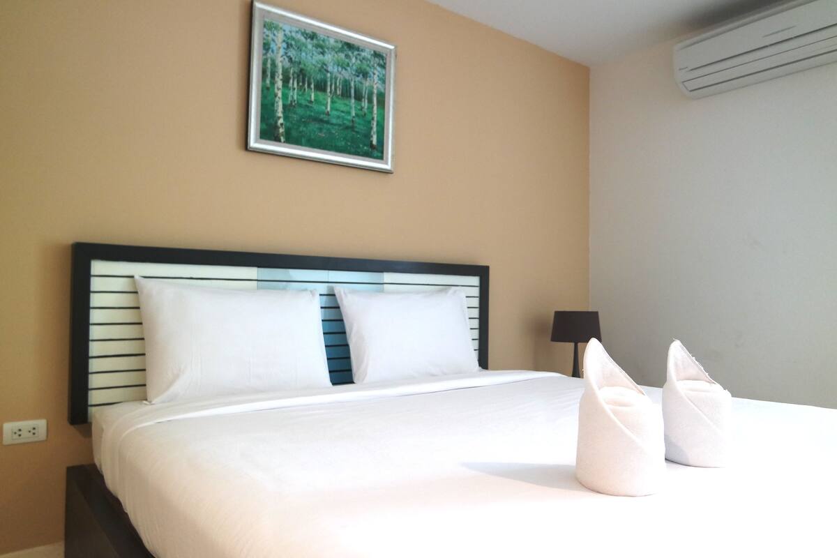 A neatly made bed with white linens and pillows, a framed picture on the wall, and a black lamp beside the bed.