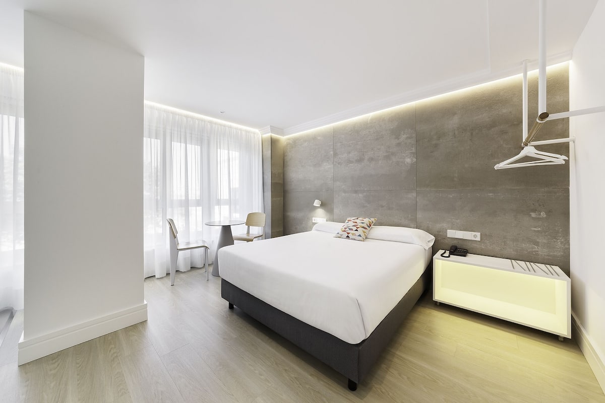 Quarto de hotel moderno com uma cama grande, mesa de cabeceira e um banco embutido.