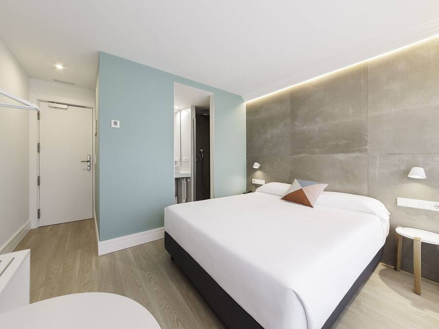 Una habitación de hotel moderna con una cama grande, una mesita de noche, una pequeña mesa redonda, un área de ducha y una puerta que da a otra habitación.