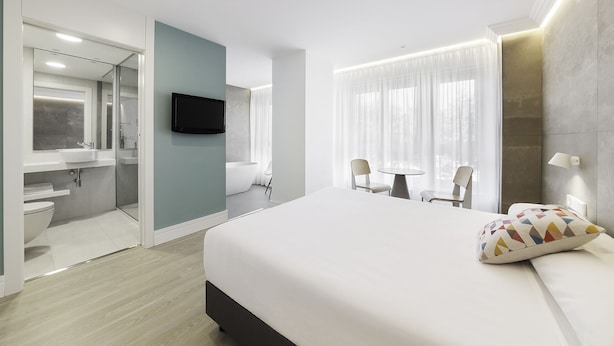 A modern hotel room with a large bed, a bathroom with a bathtub, and a dining area with chairs and a table. - Hotel Mar del Plata (A Coruña)