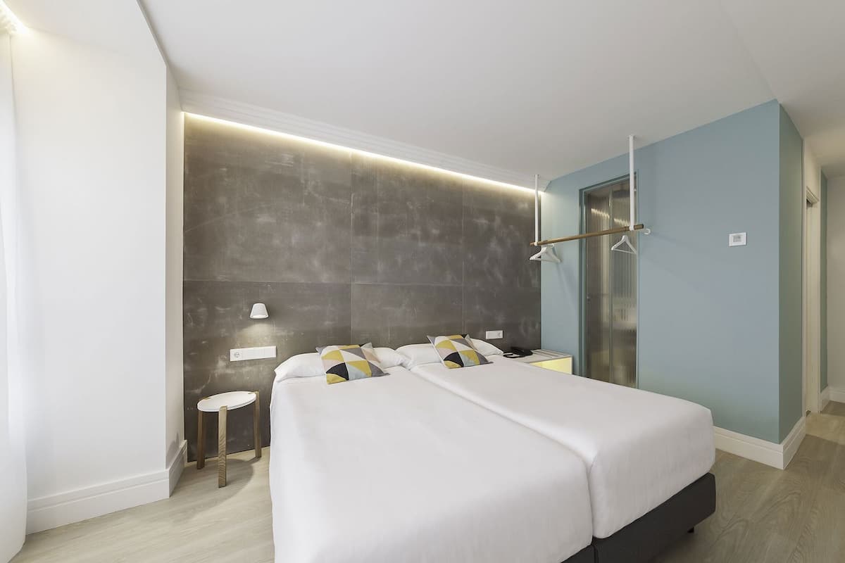 Quarto de hotel moderno com cama grande, mesas de cabeceira, luminária acoplada à parede e box de vidro.