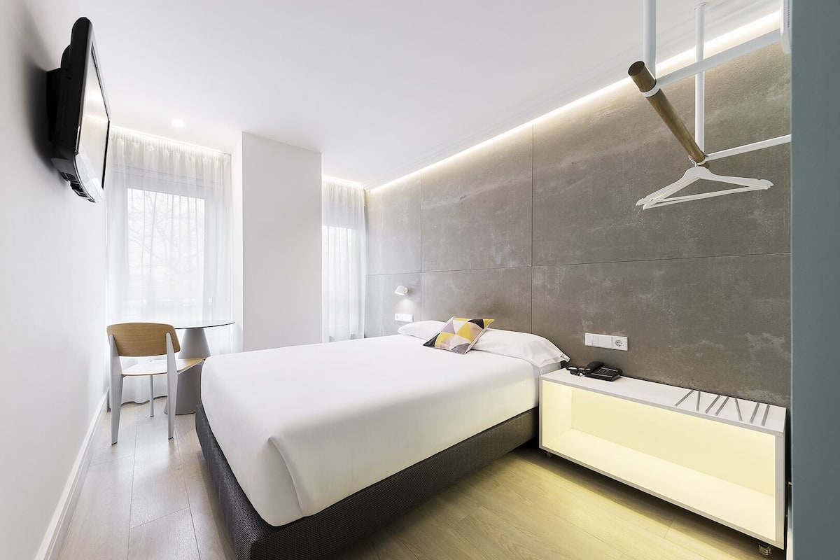 Quarto de hotel moderno com cama, mesa de cabeceira e televisão na parede.
