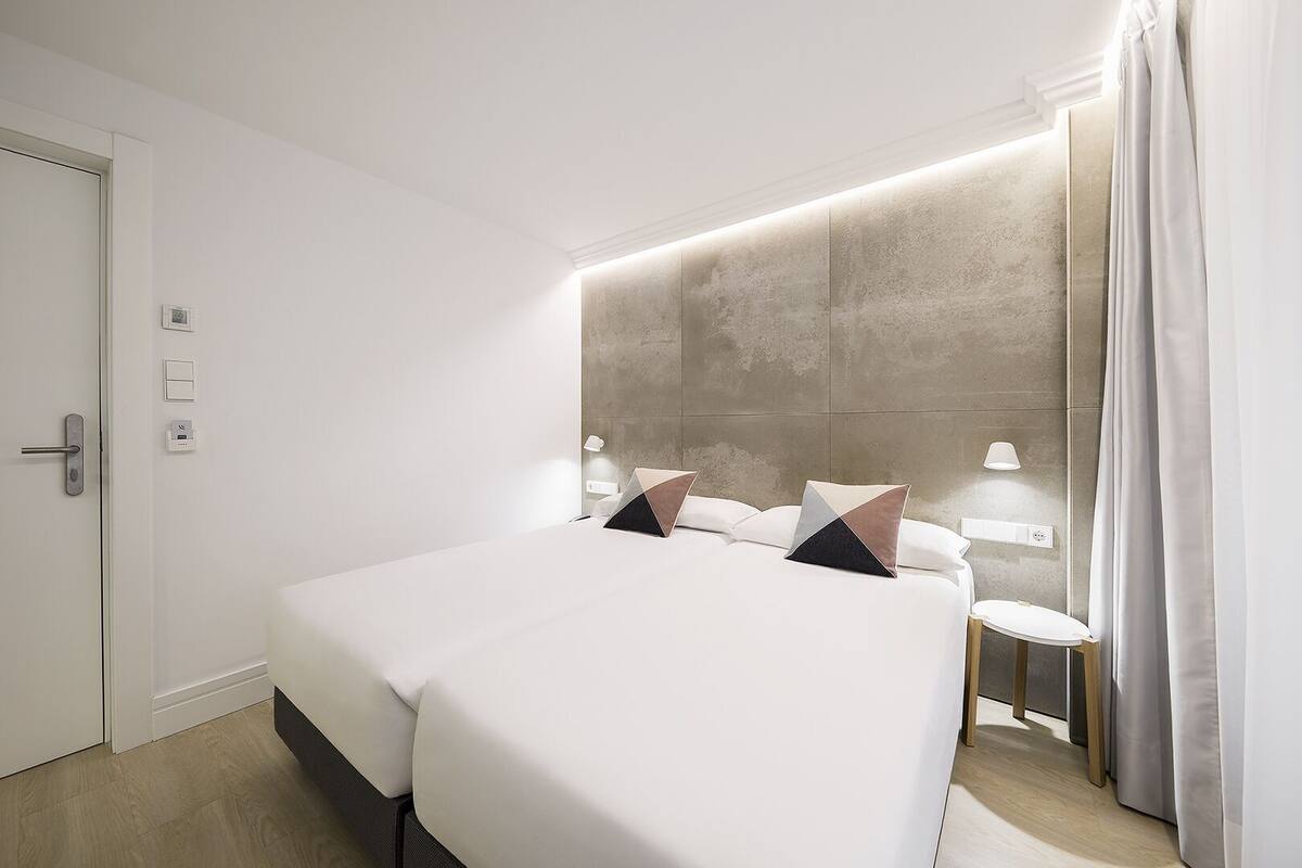 Quarto de hotel moderno com uma cama grande, abajures, uma pequena mesa de cabeceira e uma porta com puxador.