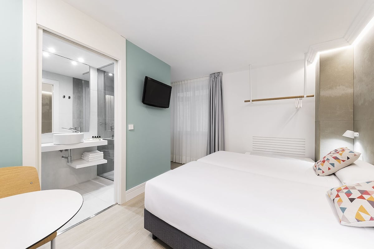 Quarto de hotel moderno com uma cama grande, TV fixada na parede, closet espaçoso e banheiro com espelho e pia.