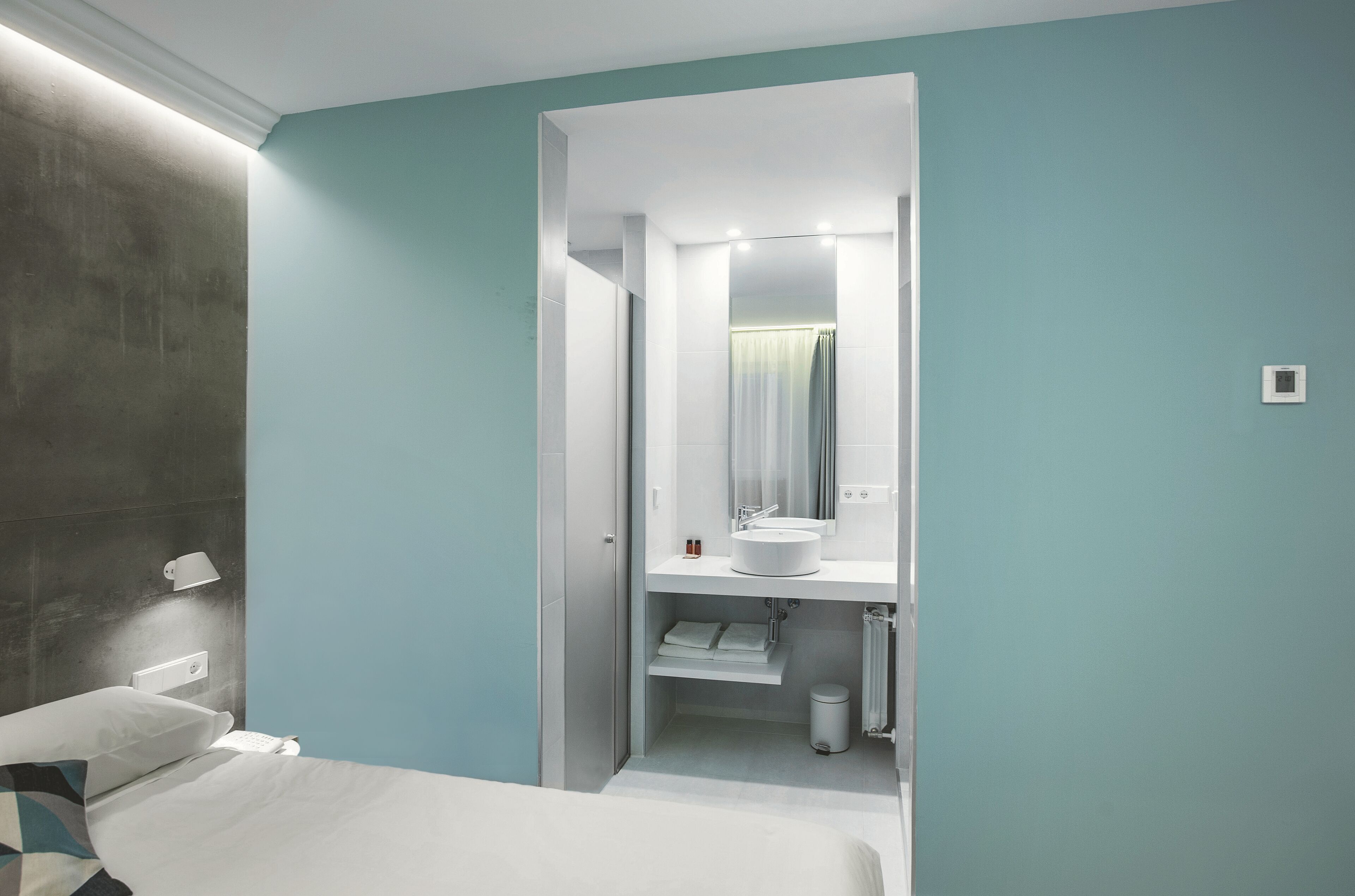 A modern hotel room with a bed, a bedside lamp, a white bathroom with a sink and mirror, and a trash bin.