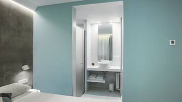 A modern hotel room with a bed, a bedside lamp, a white bathroom with a sink and mirror, and a trash bin.