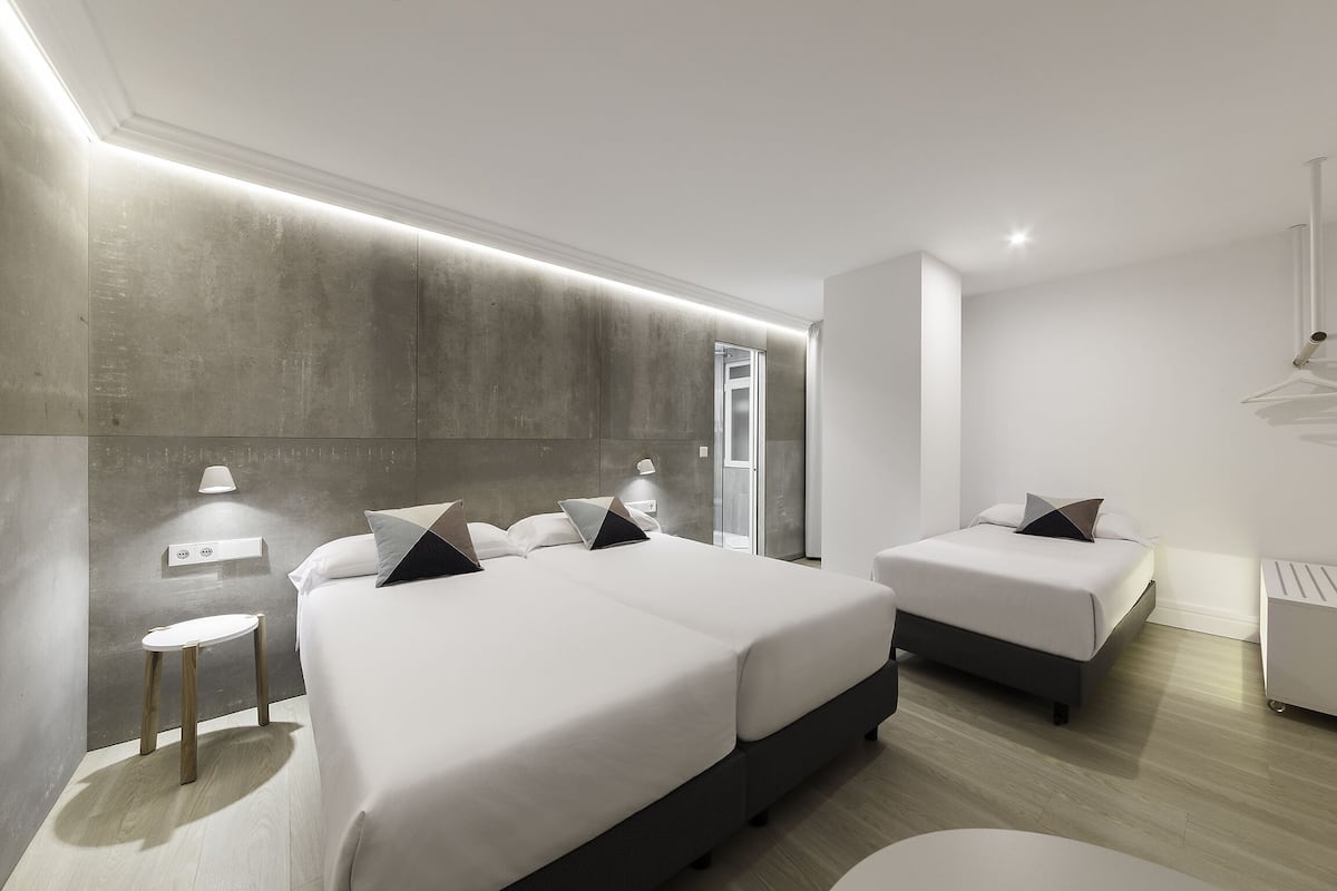 Quarto de hotel moderno com uma cama grande, uma cama menor, uma mesinha de cabeceira e um ponto de luz fixado na parede.