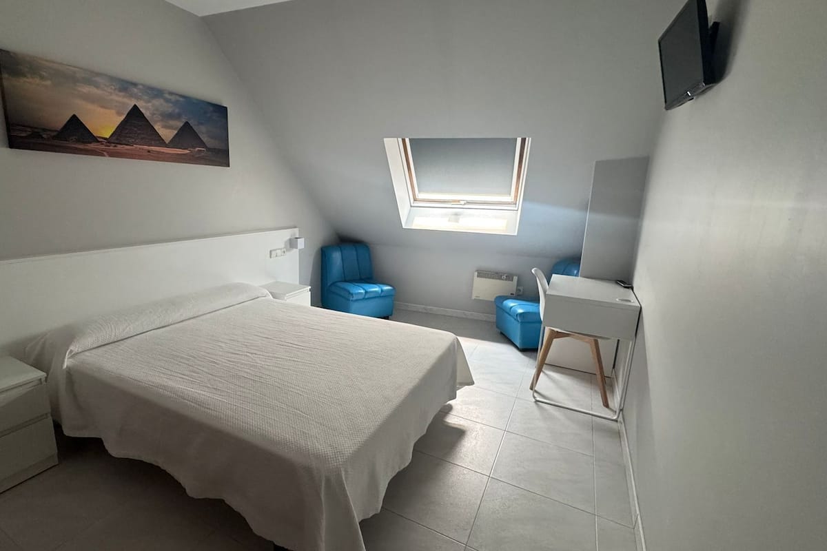 Un dormitorio con cama, escritorio, silla, ventana y un cuadro de pirámides.