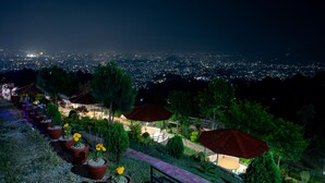 A nighttime view of a landscaped area with potted plants and umbrellas, overlooking a cityscape. - Naikap Village Resort (Chandragiri)