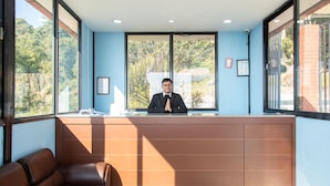 A reception area with a receptionist, a brown leather sofa, and large windows offering a view of greenery outside. - Naikap Village Resort (Chandragiri)