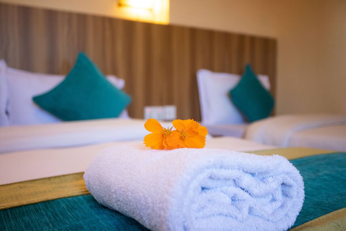 A neatly made bed with a rolled white towel and a single orange flower on the bedspread.