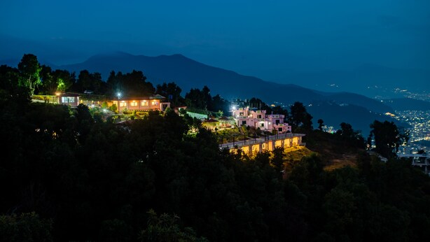 A hillside village with illuminated buildings, surrounded by trees and mountains in the background. - Naikap Village Resort (Chandragiri)