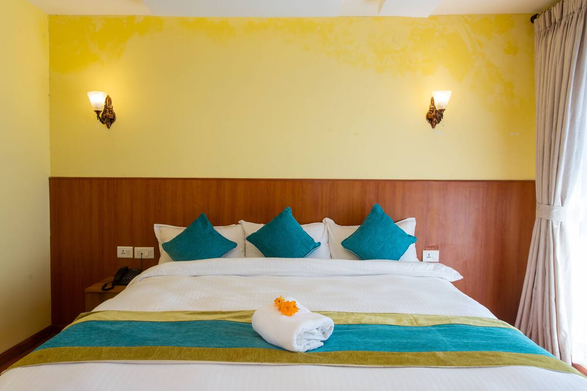 A hotel room with a wooden headboard, a bed with white bedding and teal pillows, a folded towel with an orange flower, and two wall-mounted lights.