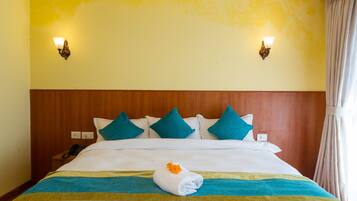 Habitación de hotel con cabecera de madera, cama con ropa blanca y almohadas color turquesa, una toalla doblada con una flor naranja y dos lámparas fijadas a la pared.