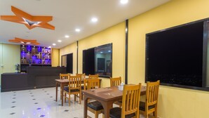 A restaurant with a yellow wall, wooden chairs, a table, and a bar area with bottles. - Naikap Village Resort (Chandragiri)