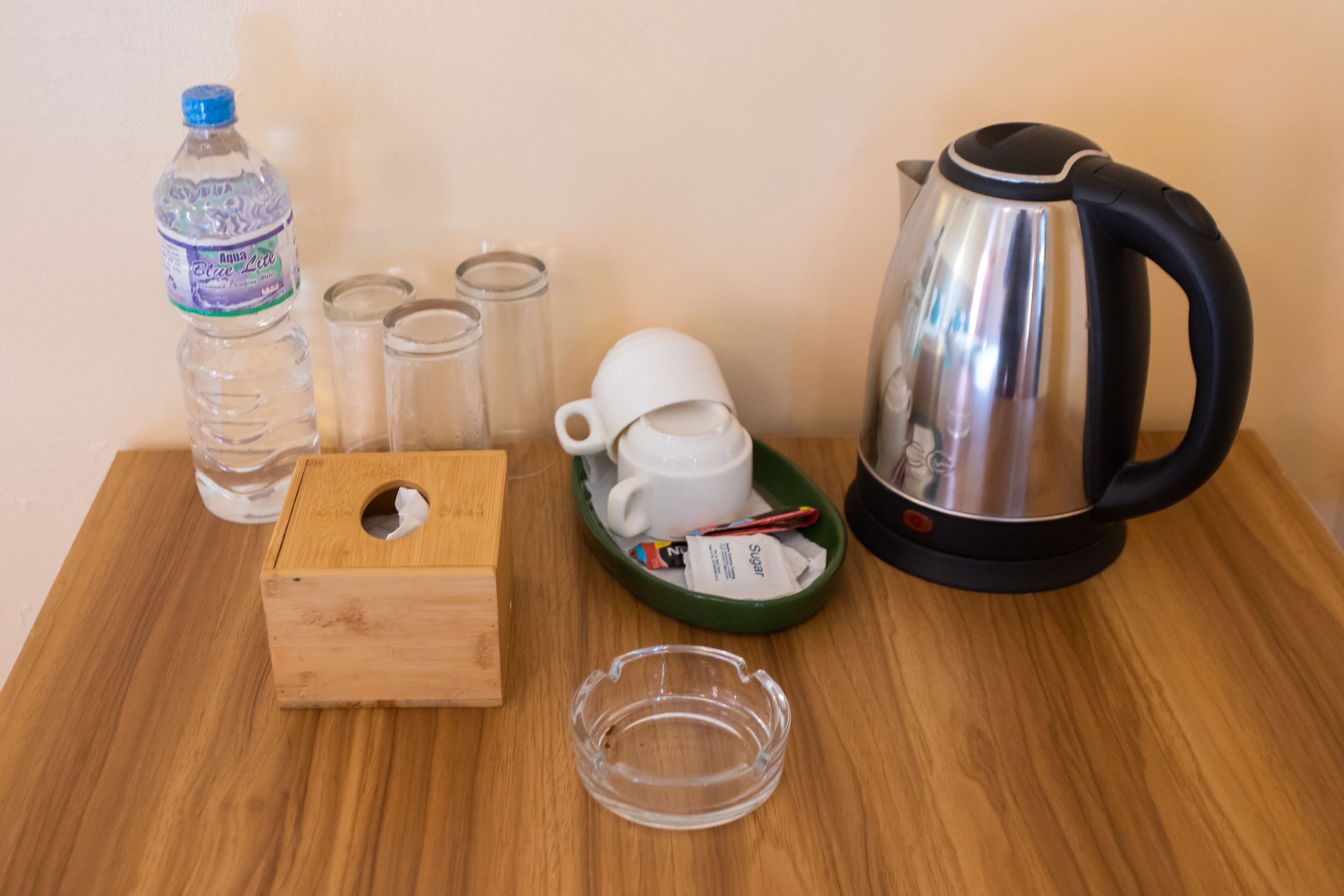 A wooden table with a water bottle, four glasses, a white mug, a sugar packet, a glass ashtray, and a kettle.
