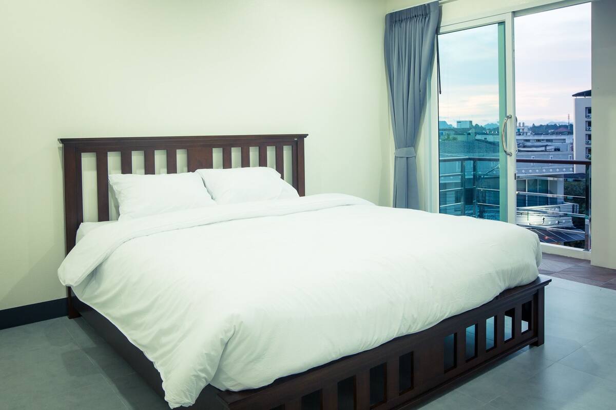 A bedroom with a wooden bed, a large window with a view of buildings, and a balcony.