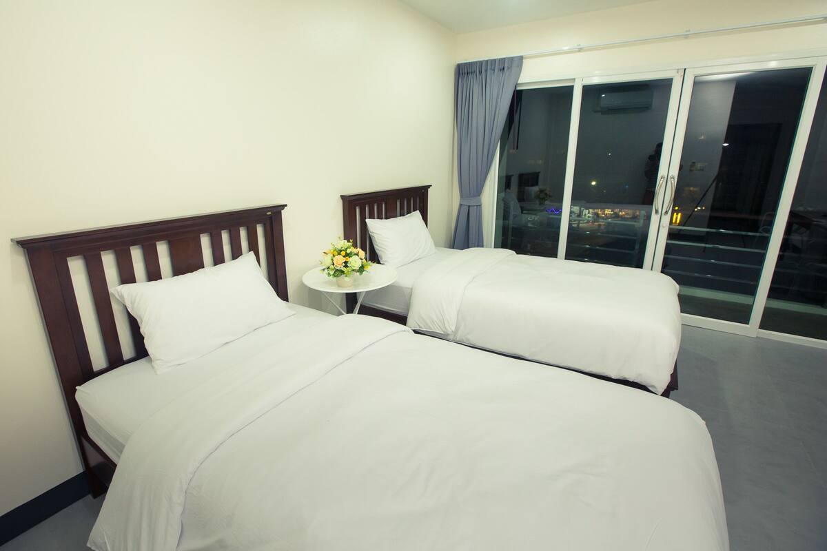 A hotel room with two beds, a small table with a vase of flowers, and a view of the city through large windows.