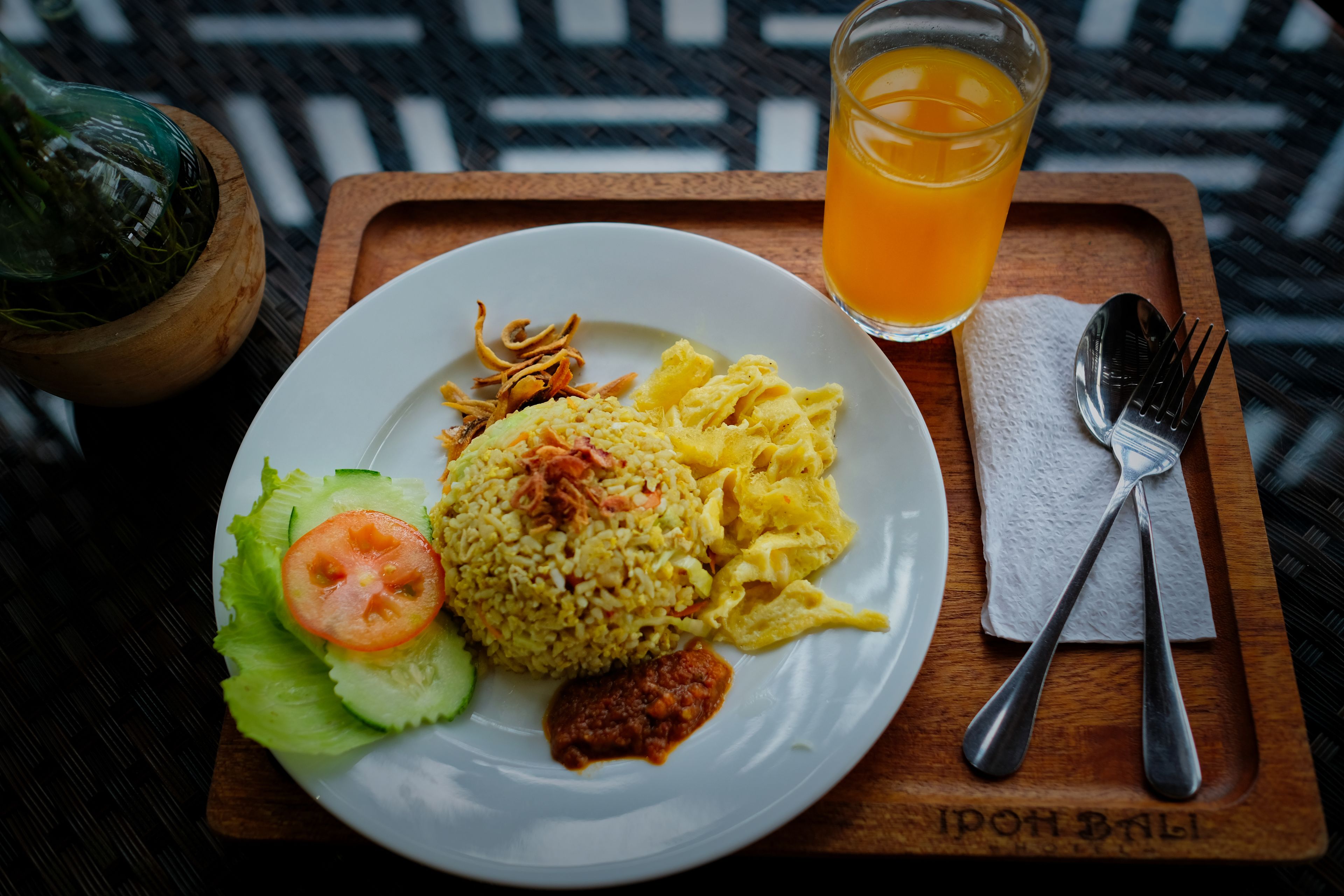 A plate of food with rice, eggs, and vegetables, a glass of orange juice, and a fork and spoon on a wooden tray.