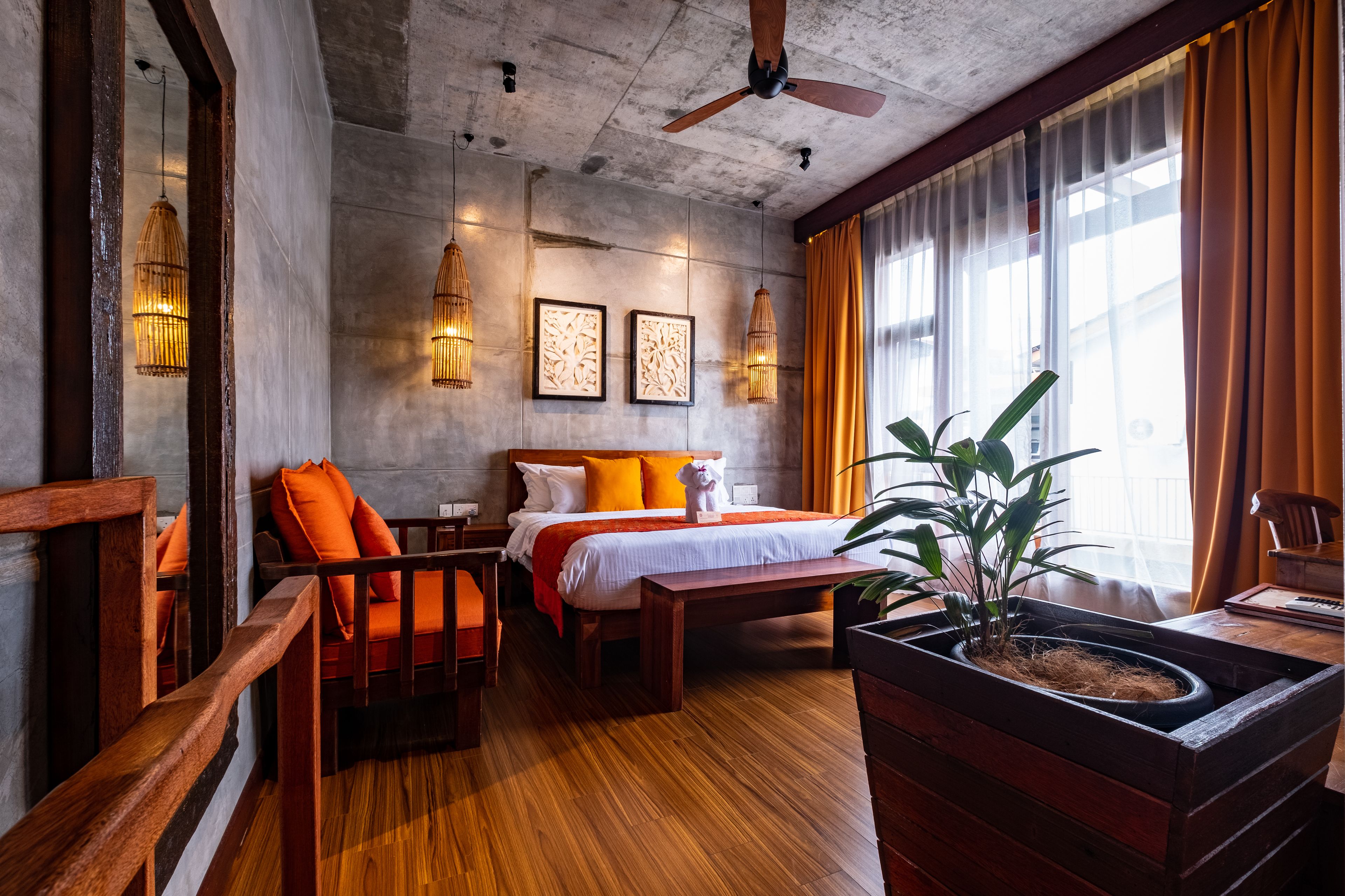 A hotel room with a wooden bed, orange pillows, a wooden bench, a potted plant, and a ceiling fan.
