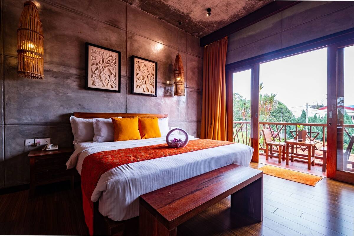 A bedroom with a large bed, wooden headboard, a bench, a nightstand, a bedside table, a wall-mounted lamp, and a view of the outdoors.