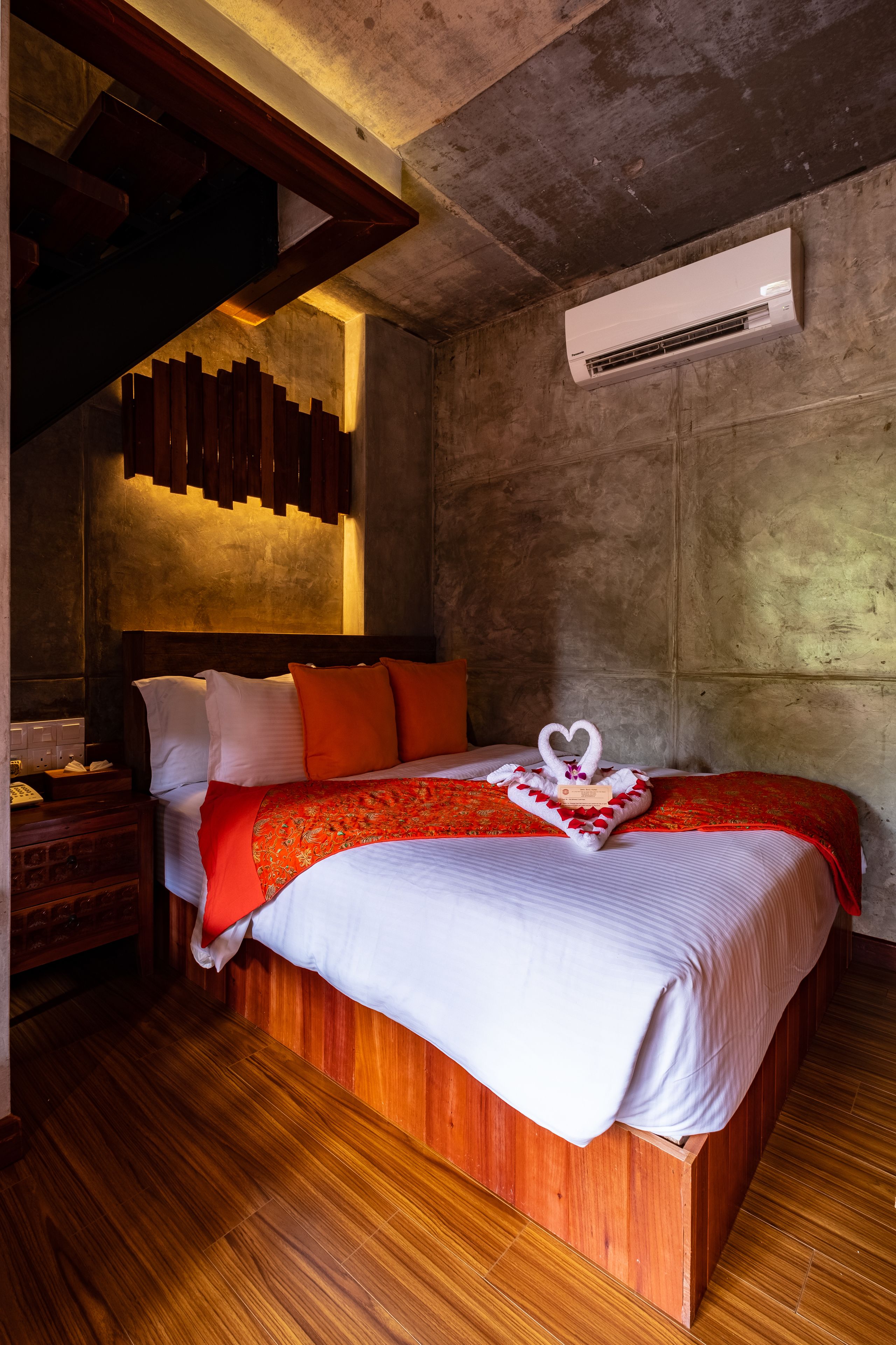 A modern hotel room with a large bed, wooden flooring, a built-in headboard, and an air conditioning unit on the wall.