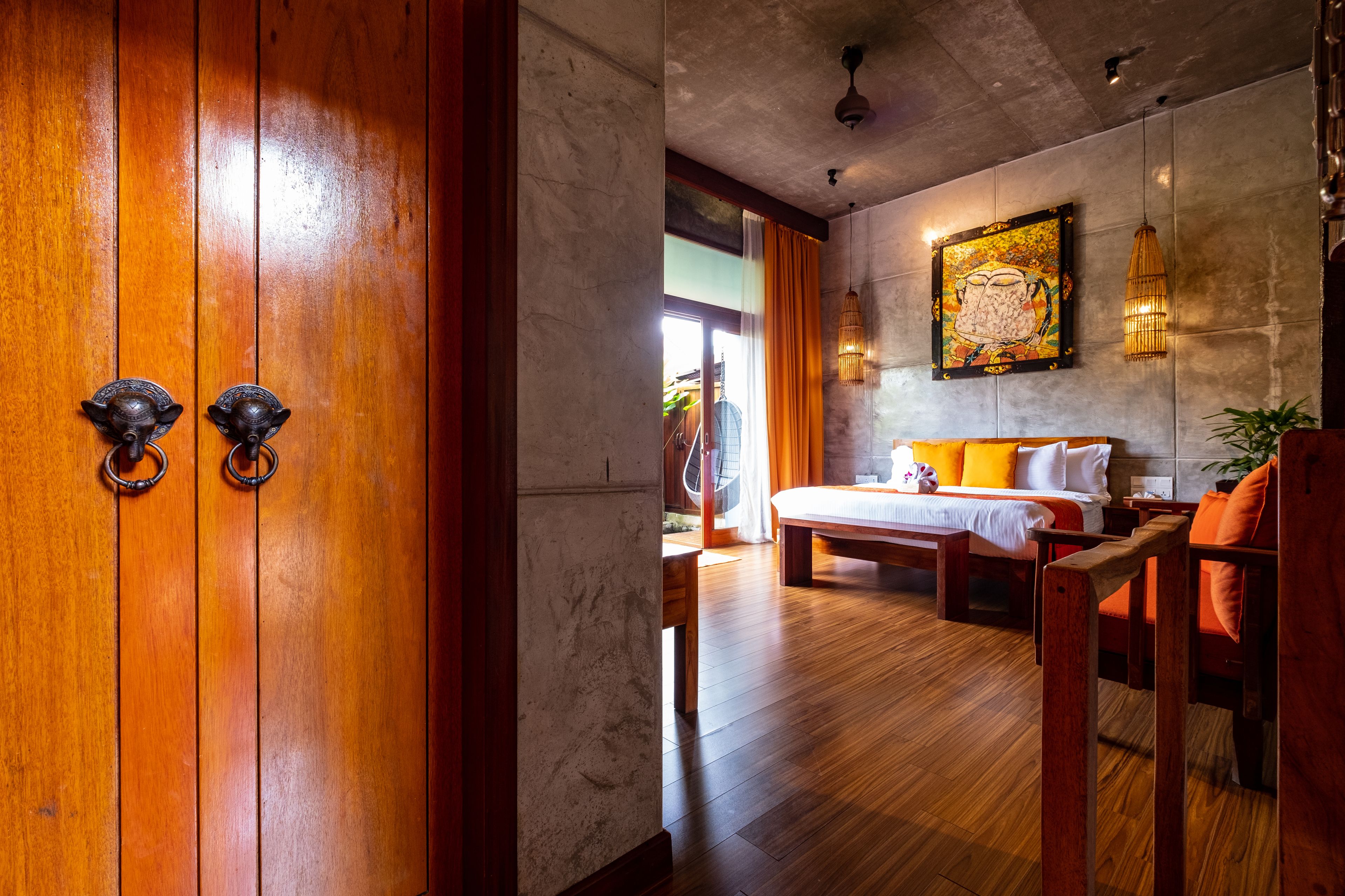 A hotel room with a wooden door, a bed with orange bedding, a large painting on the wall, and a hanging light fixture.