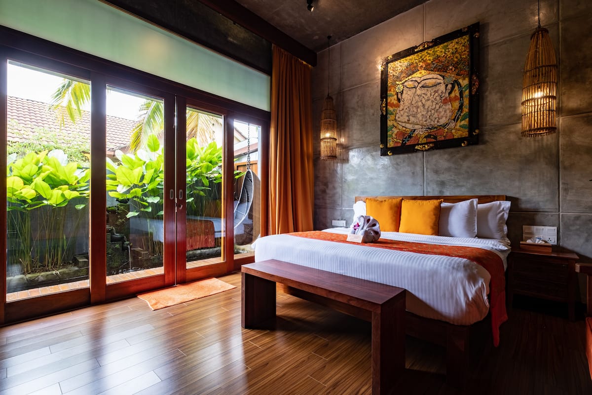 A bedroom with a large bed, wooden flooring, a bench, a nightstand, and a view of greenery through glass doors.