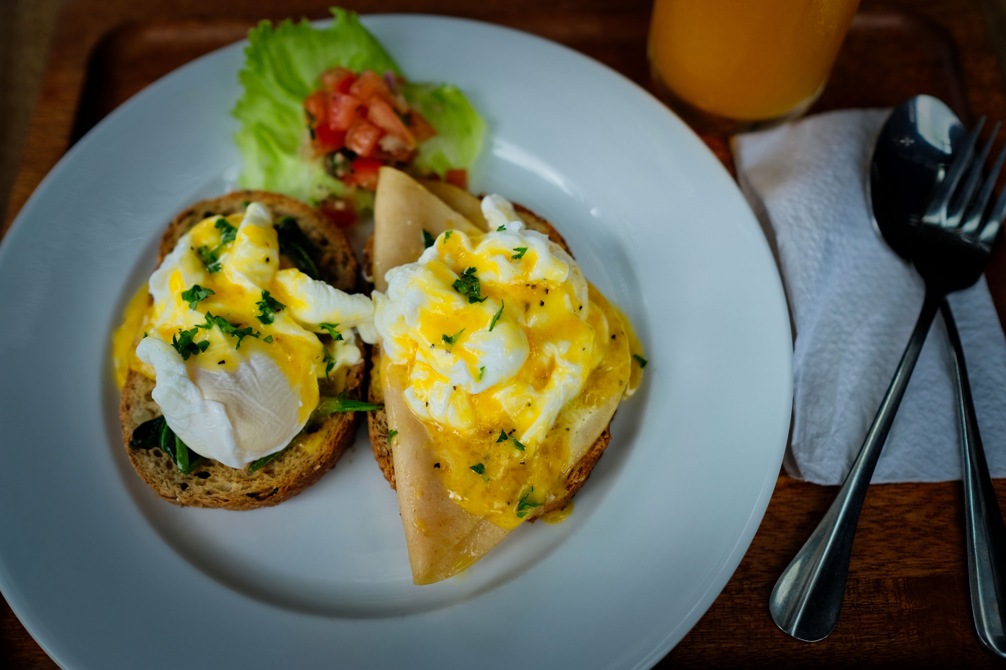 Un plato con dos sándwiches abiertos cubiertos de huevos pochados y queso, una ensalada y un vaso de jugo de naranja.