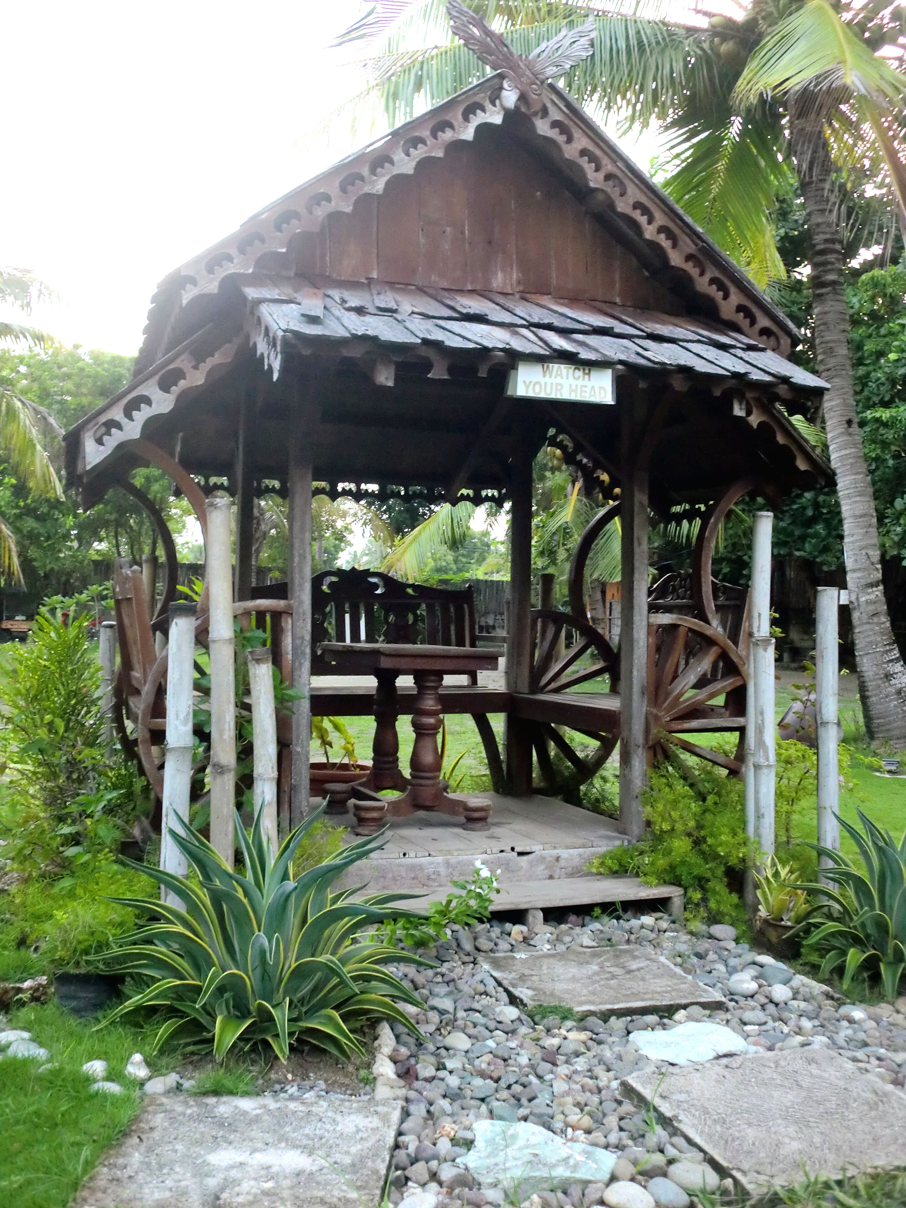 A wooden gazebo with a sign that reads "WATCH YOUR HEAD," surrounded by lush greenery and palm trees.