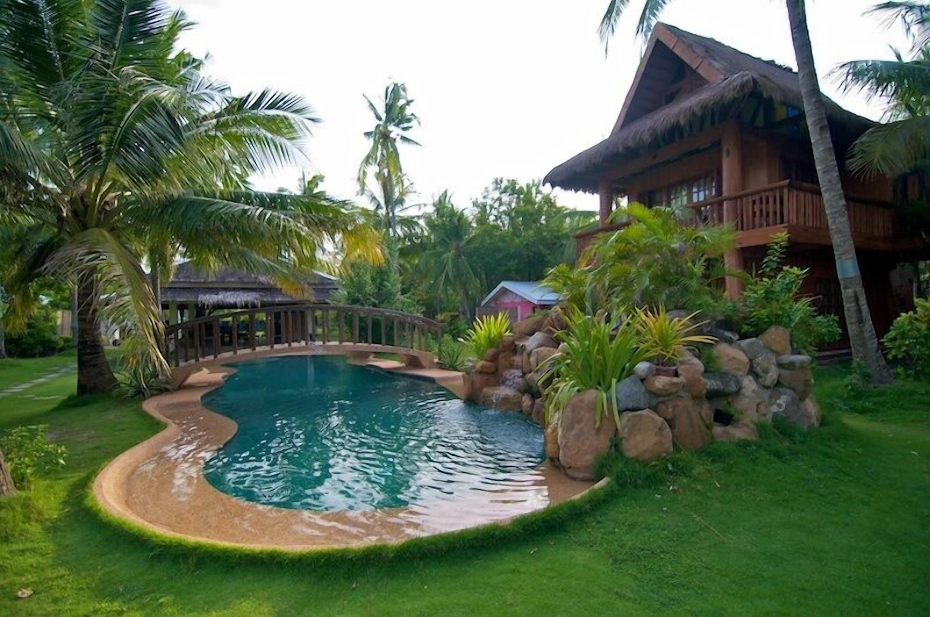 A pool surrounded by lush greenery and a wooden house with a thatched roof.