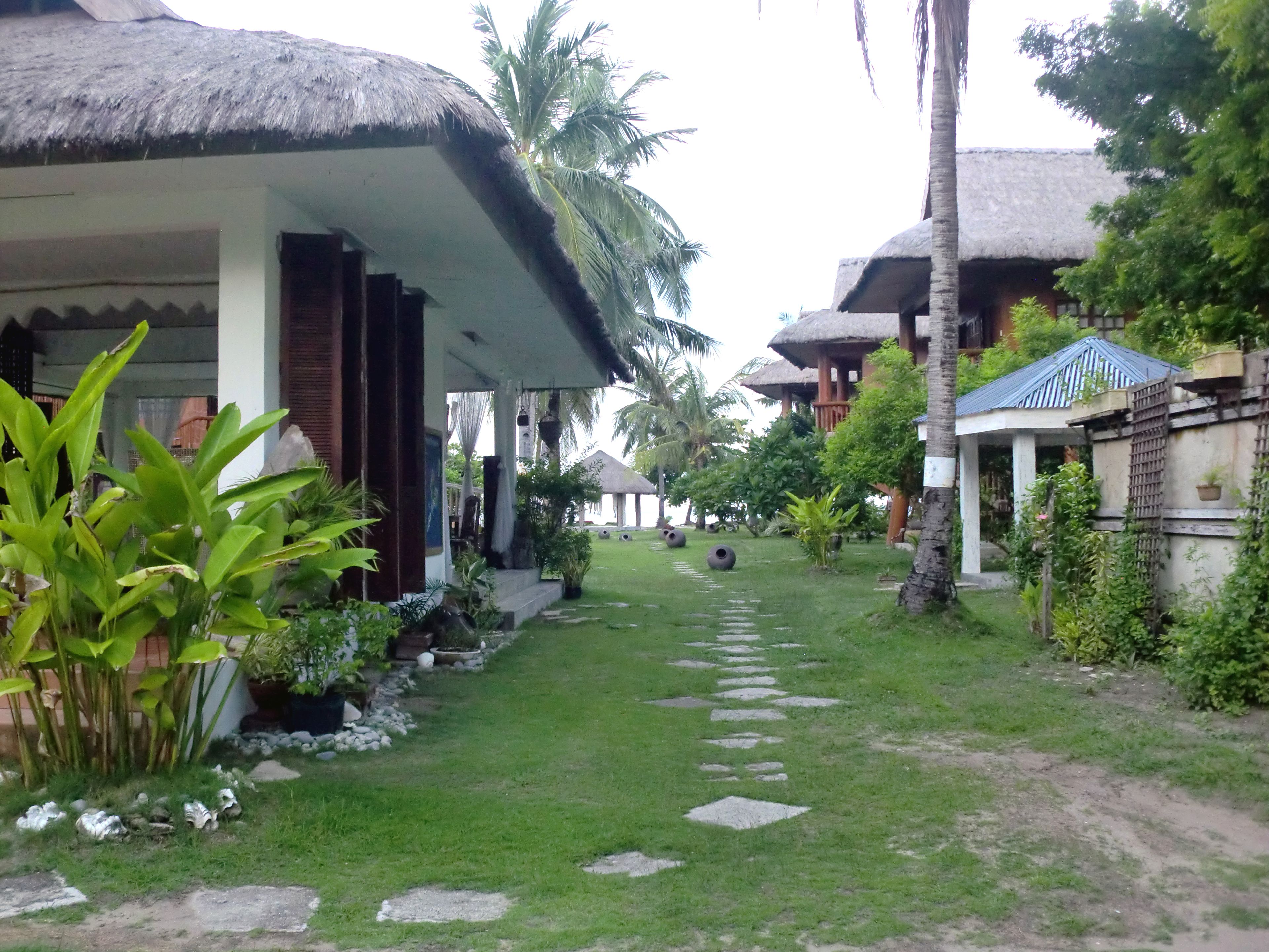 A pathway leading to a thatched-roof building with a covered porch, surrounded by lush greenery and palm trees.