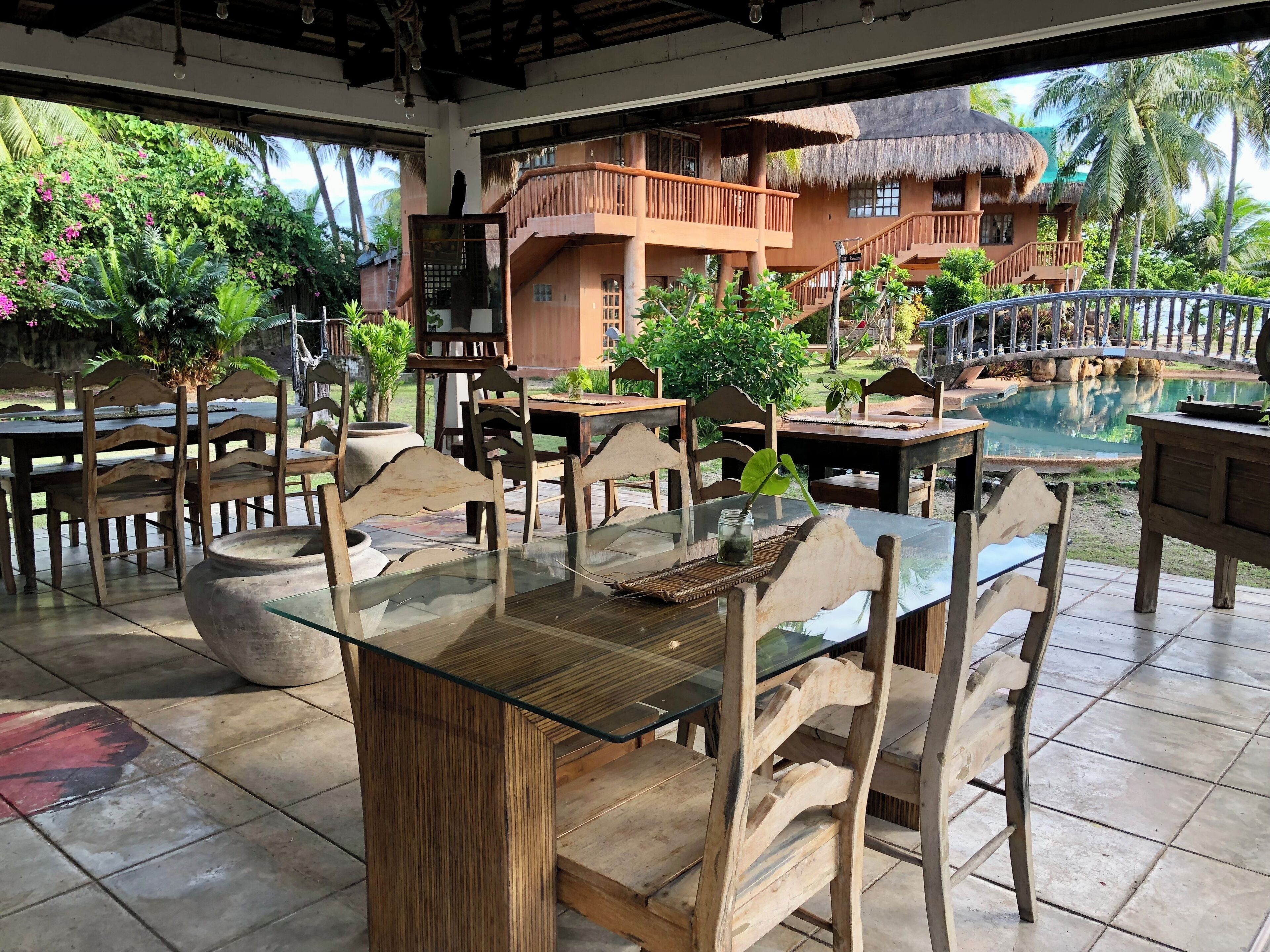 An outdoor seating area with wooden furniture and glass-top tables, surrounded by lush greenery and a view of a thatched-roof building.