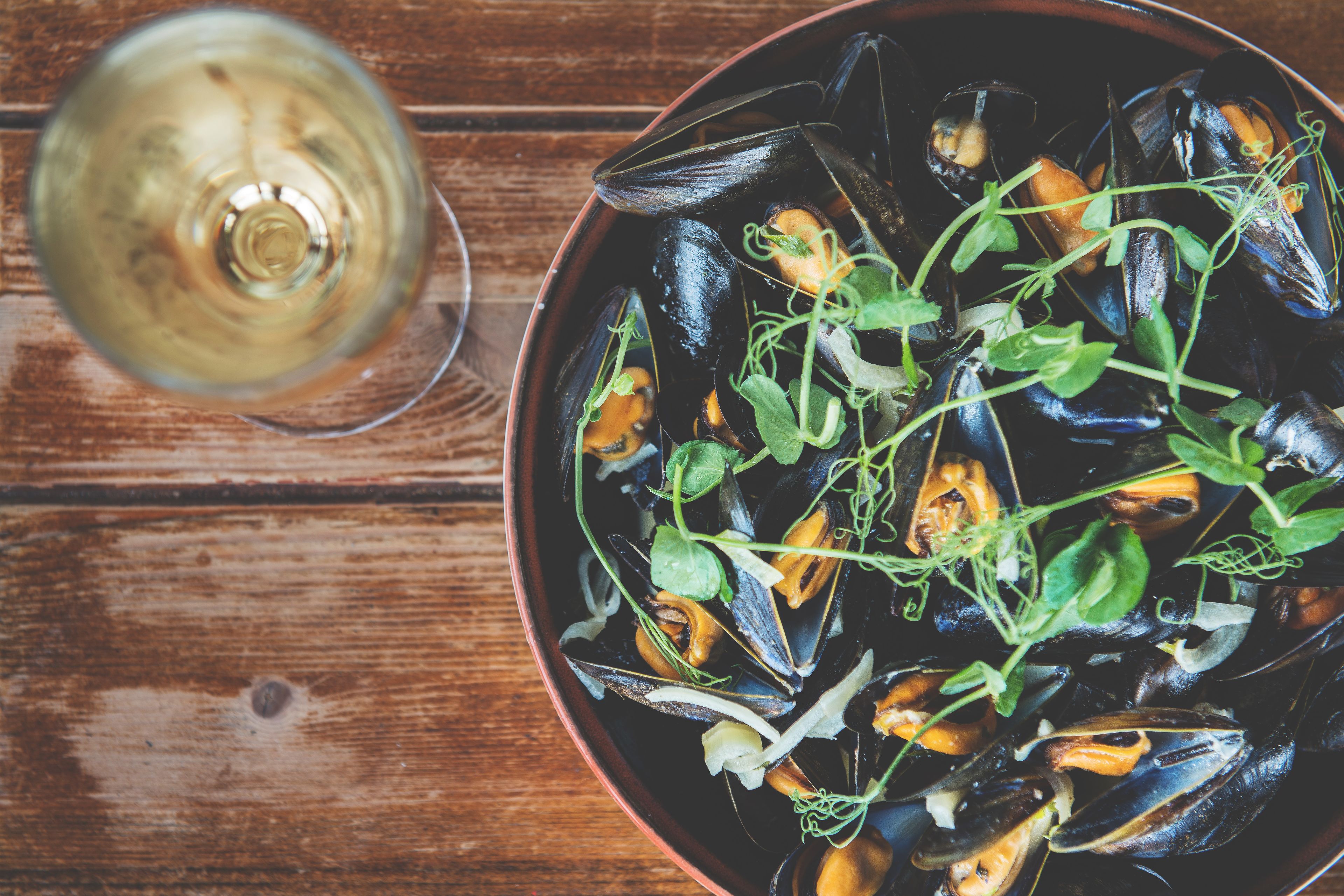 A bowl of mussels with greens on a wooden table.