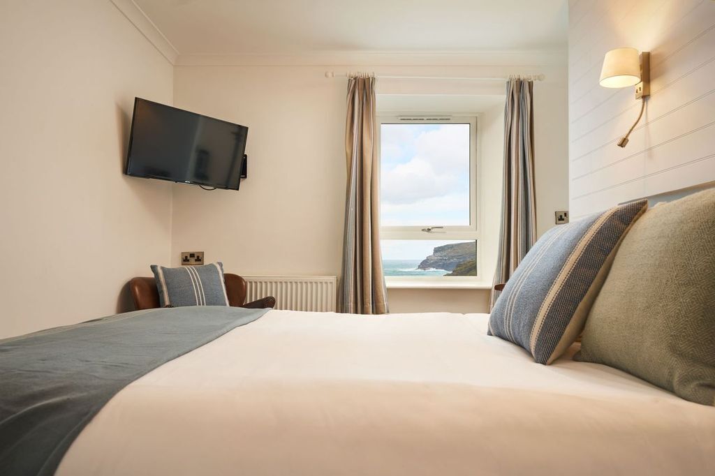Una habitación de hotel con una cama, televisión montada en la pared, ventana con cortinas y vista al mar.