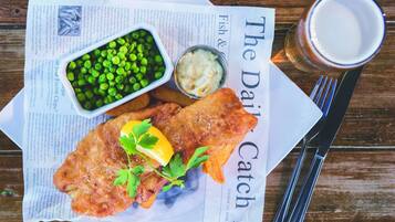 A meal of fish and chips with peas and sauce, a glass of beer, and cutlery on a newspaper.