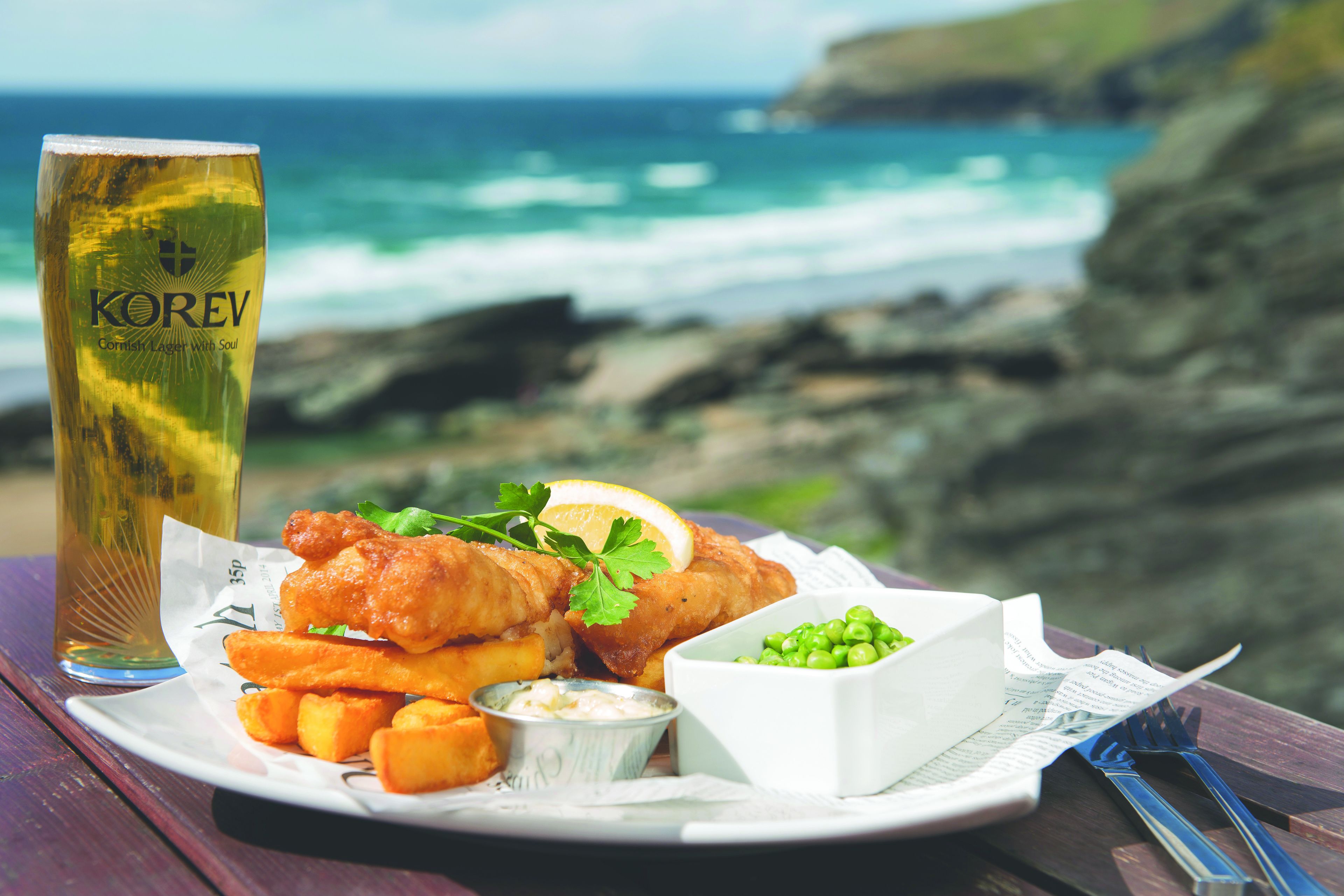 A meal of fish and chips, a glass of beer, and peas on a plate, set against a coastal backdrop.
