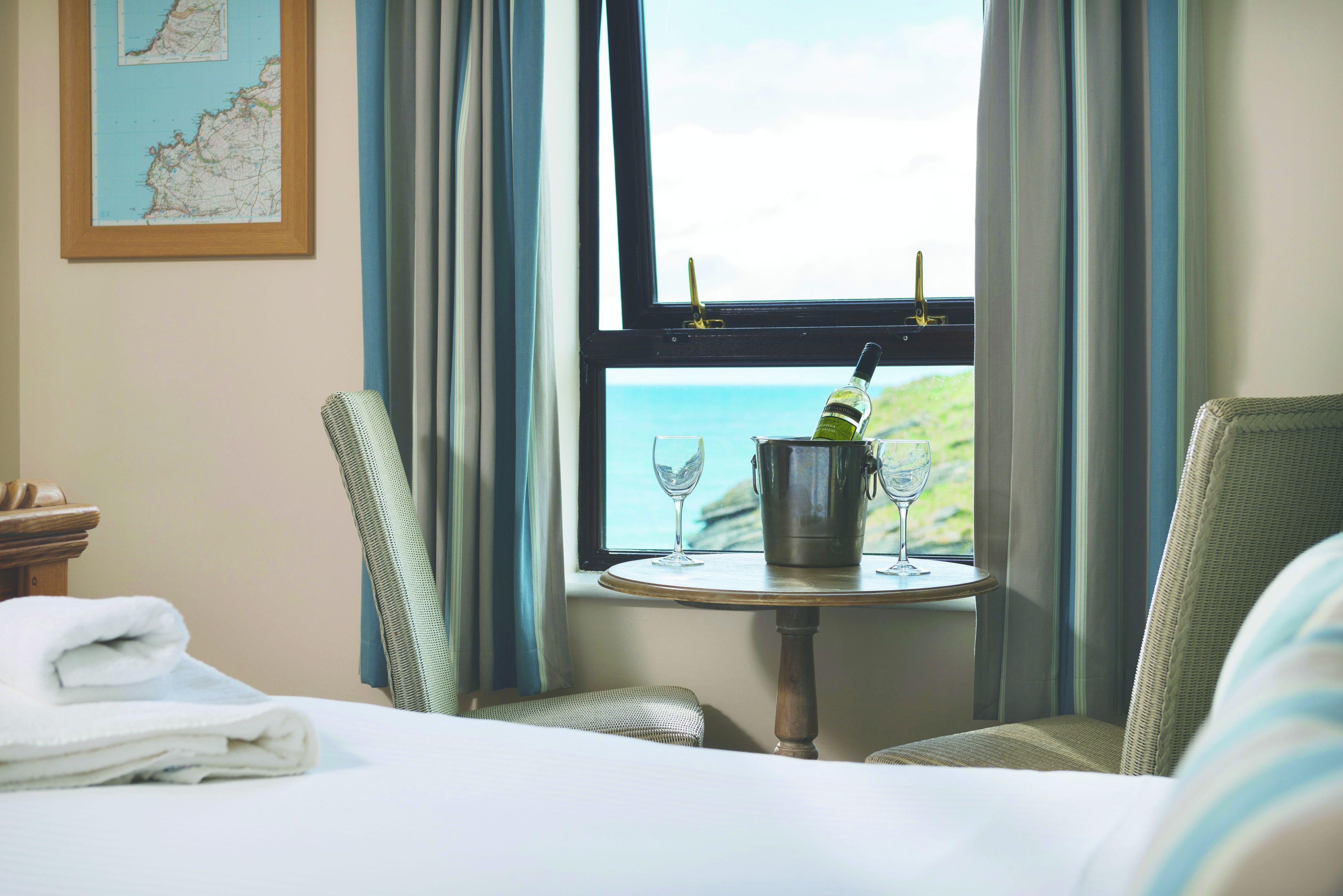 Una habitación de hotel con una cama, dos sillas, una mesita con una copa de vino y una botella, y una ventana con vistas al mar.