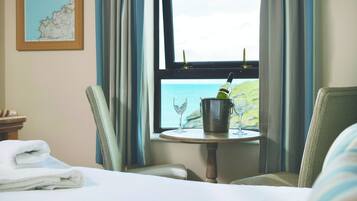 Una habitación de hotel con una cama, dos sillas, una mesita con una copa de vino y una botella, y una ventana con vistas al mar.