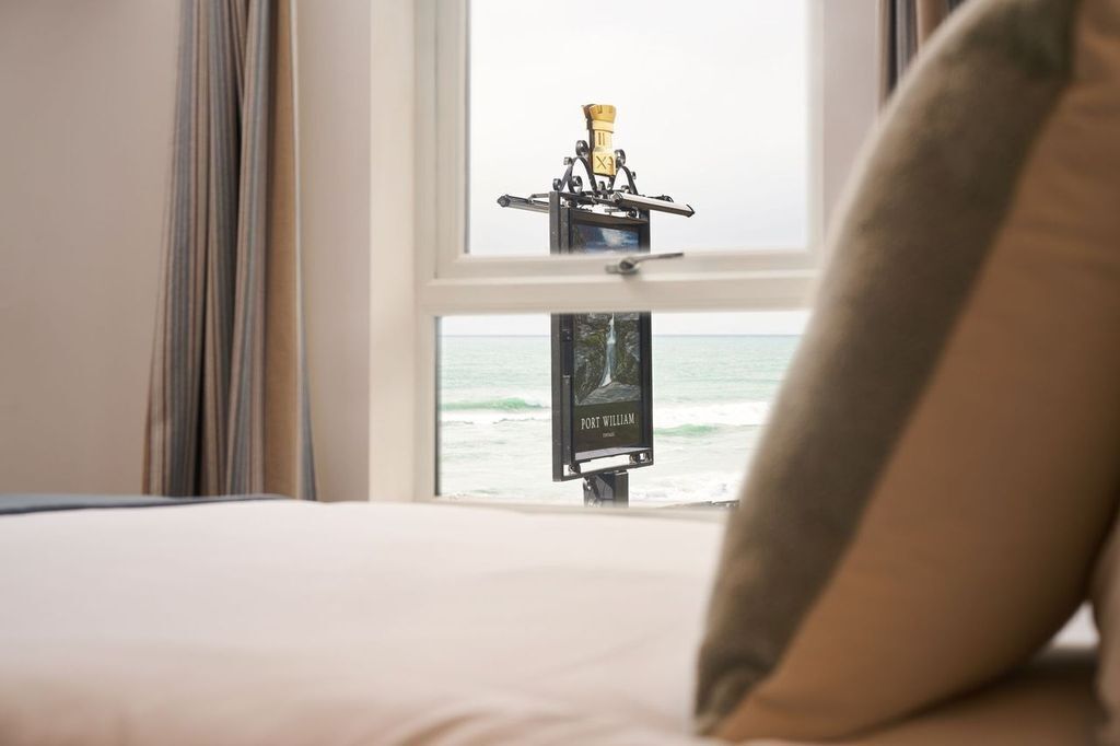 A bed with a pillow, a window with a view of the ocean, and a clock on the wall.