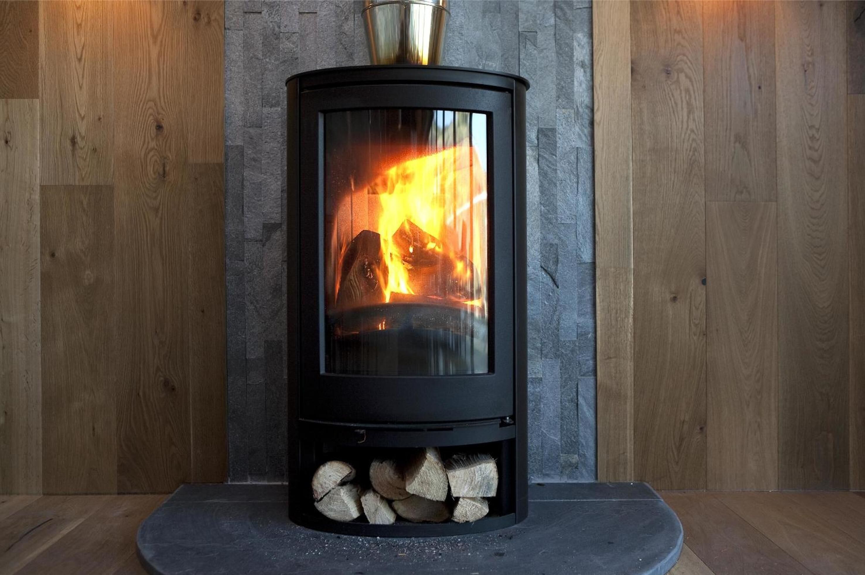A wood-burning stove with visible flames, set against a stone wall and wooden panel backdrop.
