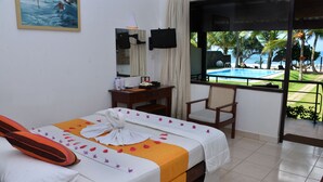 A hotel room with a bed, a desk, a chair, a window with a view of the pool and palm trees, and a painting on the wall. - Sea Rock Villa (Induruwa)