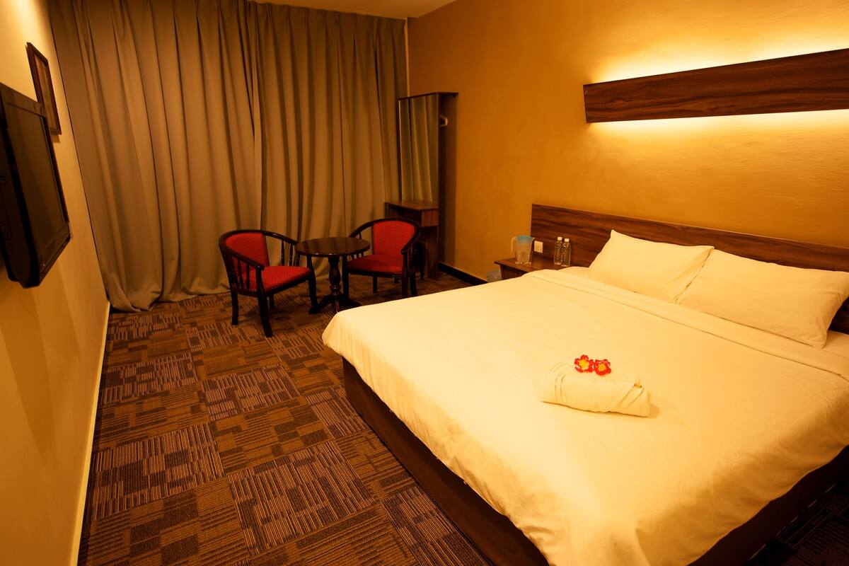 Una habitación de hotel con una cama grande, una mesita pequeña, dos sillas, una mesita de noche y una cortina.