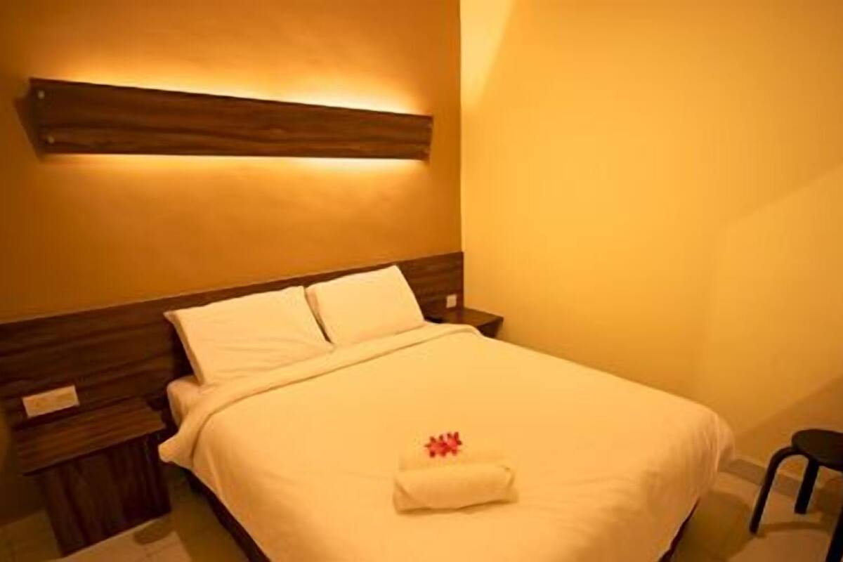 Una cama bien hecha con un edredón blanco y una toalla doblada. Se aprecia un cabecero de madera y una mesita de noche con una lámpara.