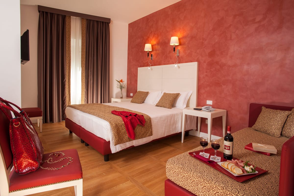 A hotel room with a large bed, a bedside table with a lamp, a small table with a wine bottle and glasses, a red handbag, and a red couch.