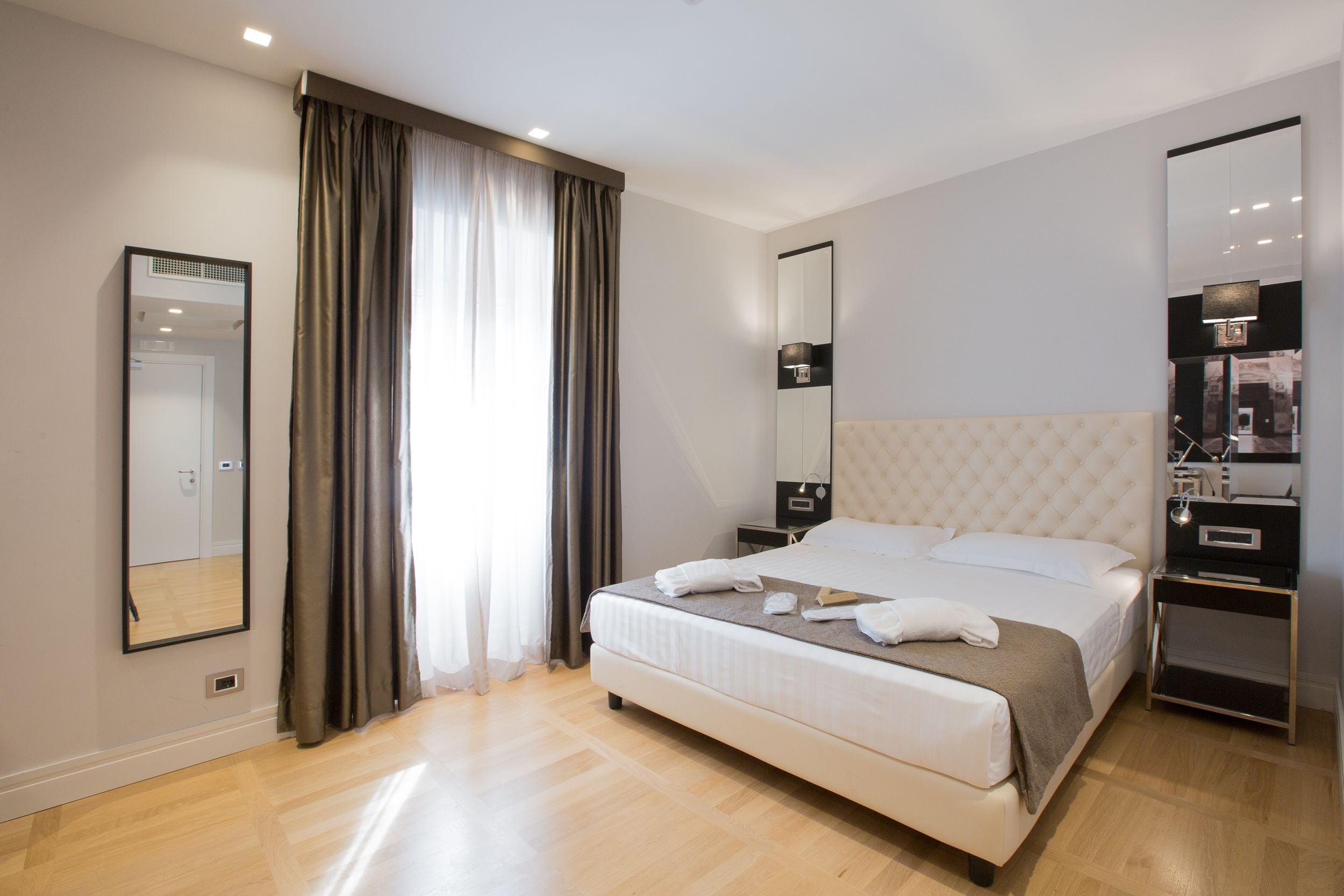 A modern hotel room with a large bed, a bedside table, a mirror, and a wardrobe.